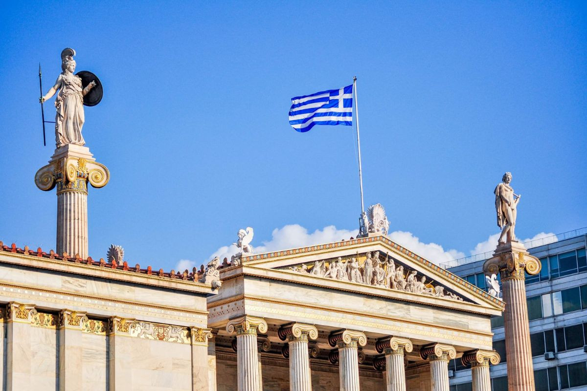 Statues atop columns flank the Academy of Athens with the Greek flag waving in the background under a clear sky.